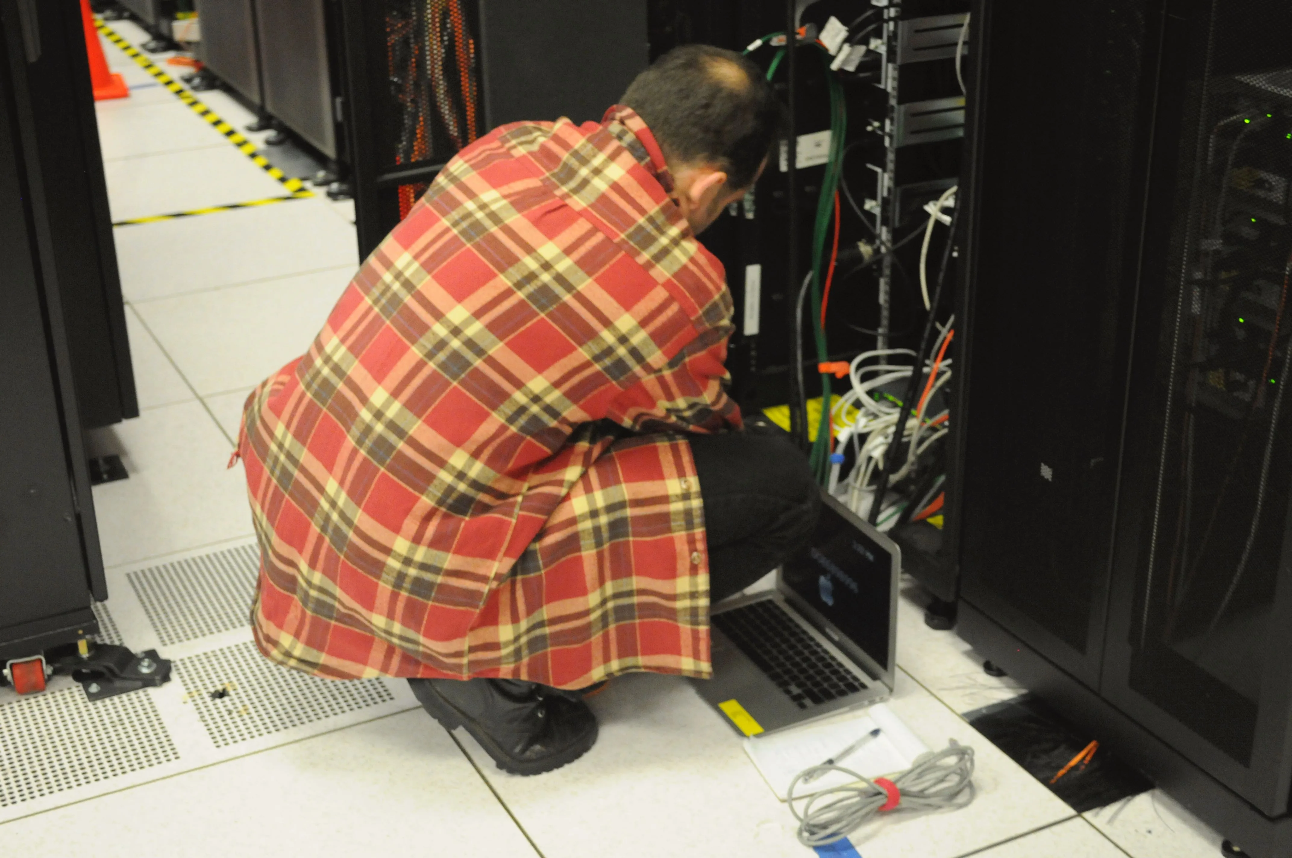 A technician kneels in front of an open server rack in a data centre. A laptop on the floor is connected to the rack with a cable, and rows of black server cabinets stand on both sides.