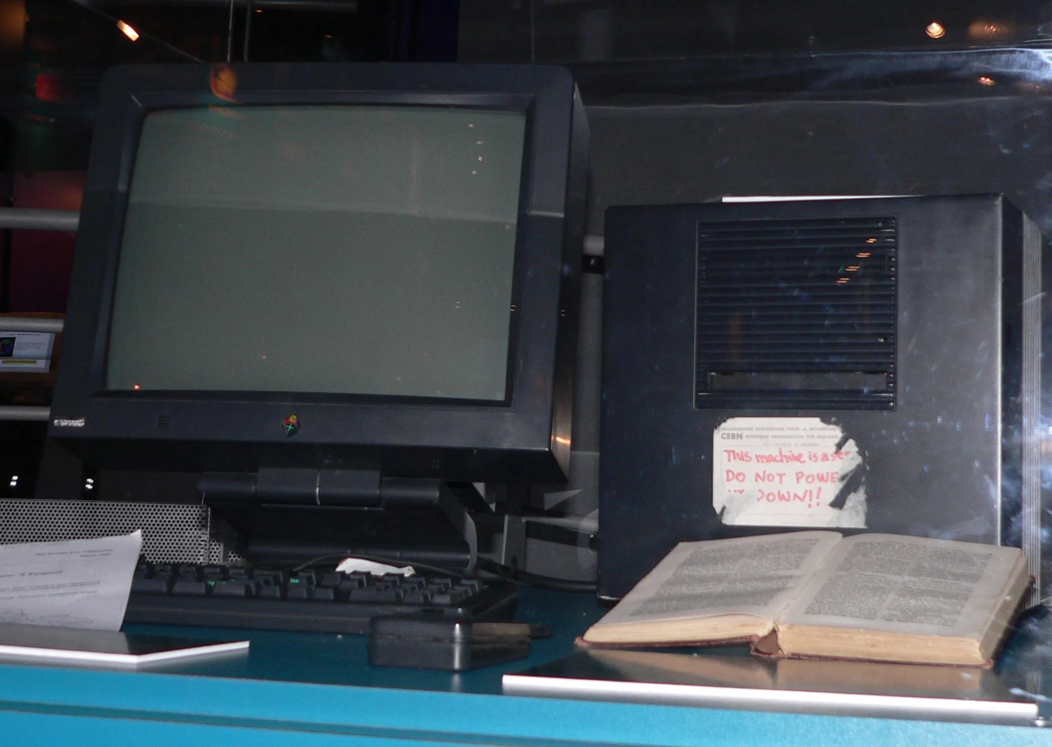 A black NeXT computer and its monitor in a museum display case. A handwritten sticker on the computer warns that this machine is a server and must not be powered down.