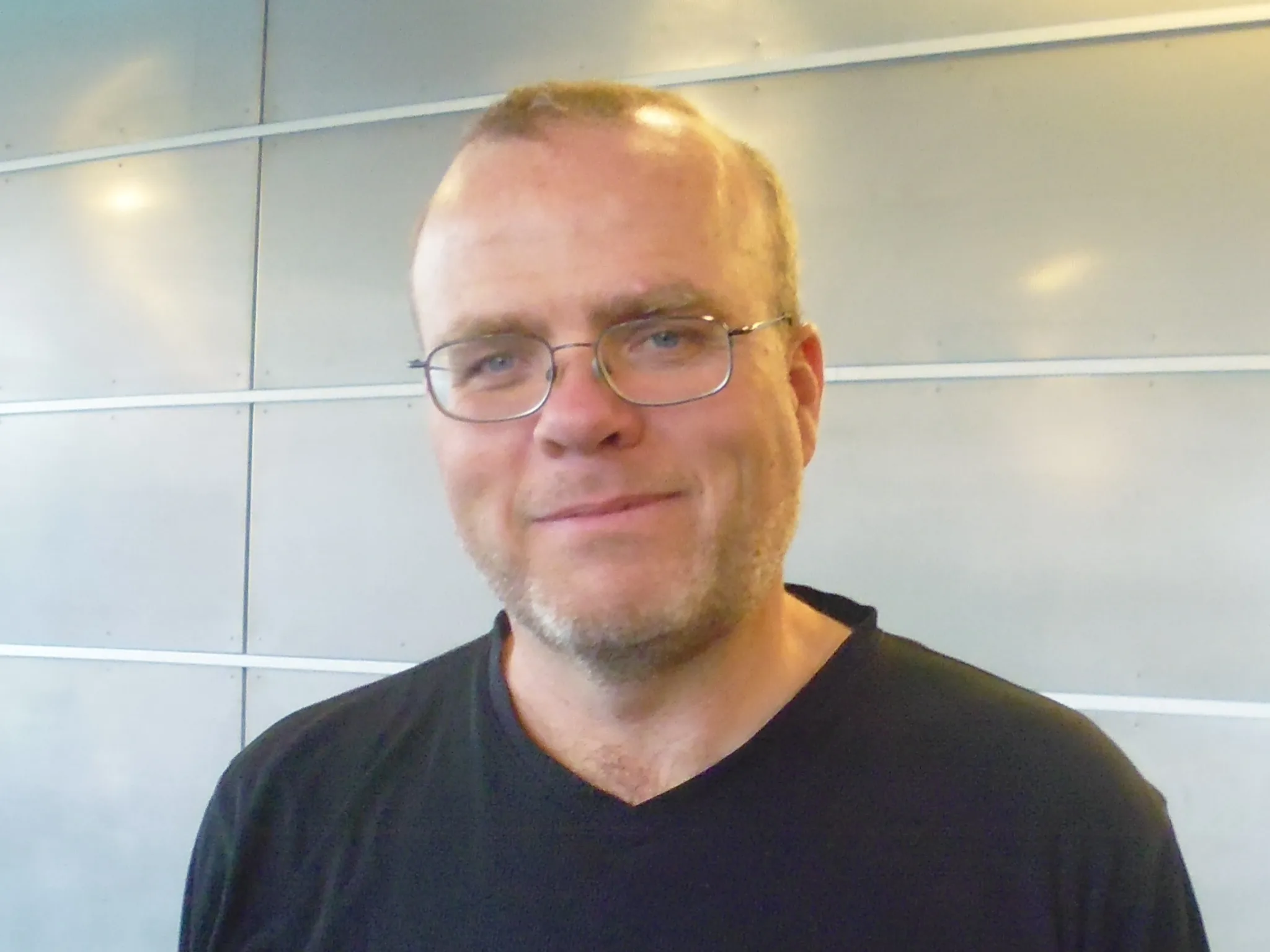 Photo of Rasmus Lerdorf, a man with short hair and glasses, wearing a dark T-shirt, in front of a light grey wall.