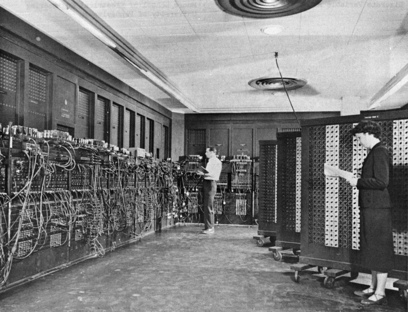Black-and-white photo: two people working at the tall panels of ENIAC, which stand along the walls of a whole room.