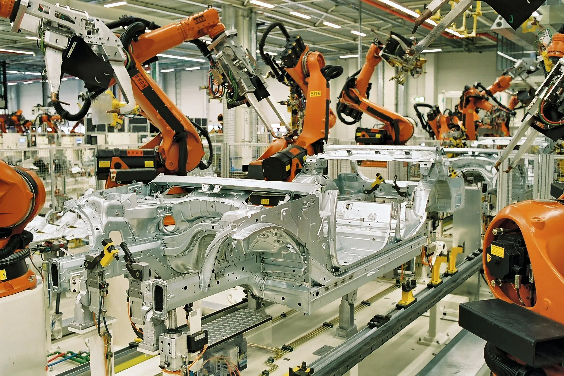 Orange industrial robot arms working on the bare metal body of a car on a factory production line.