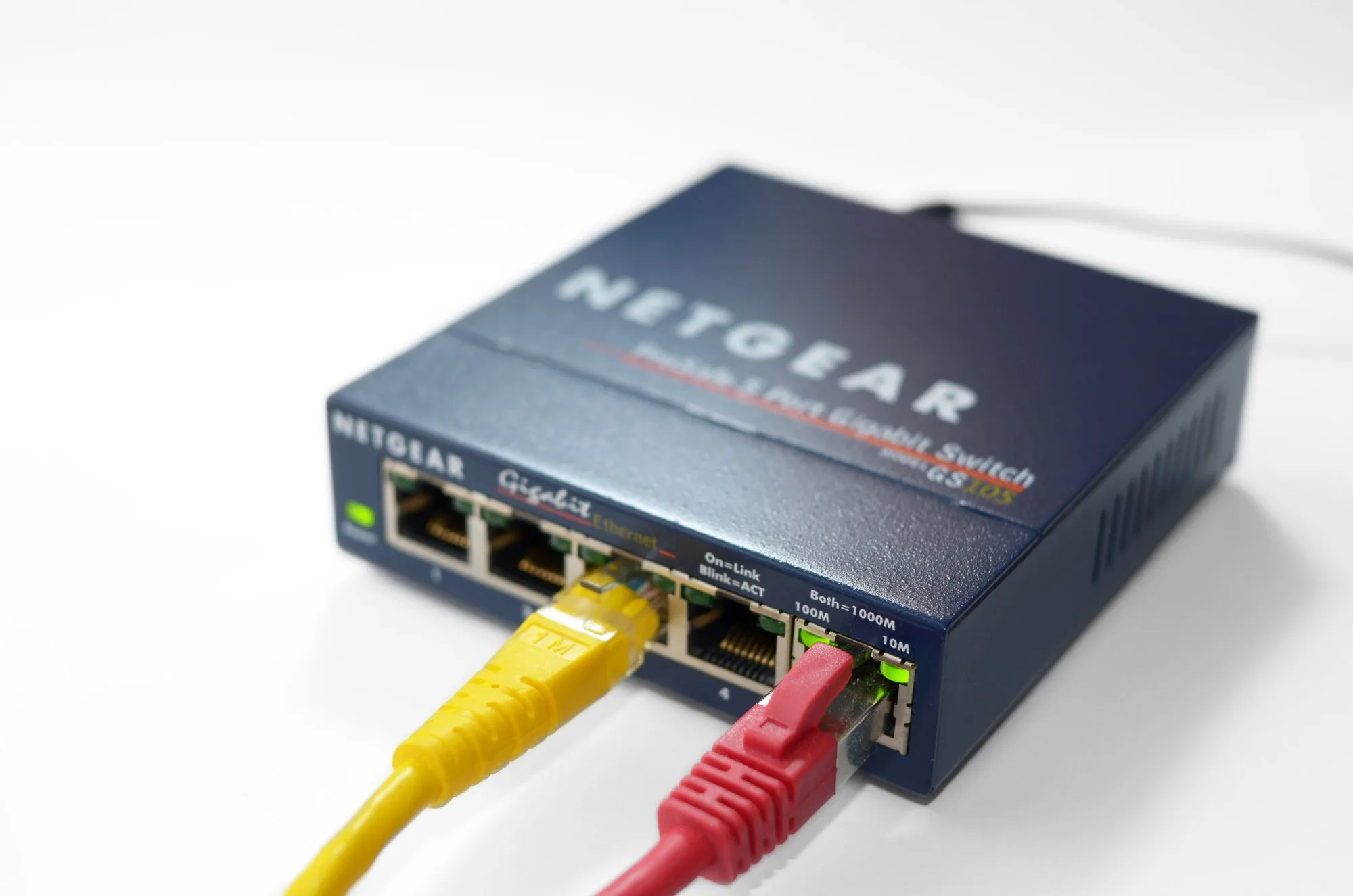 A small blue Netgear 5-port switch on a white table, with a yellow and a red network cable plugged in and green lights on.