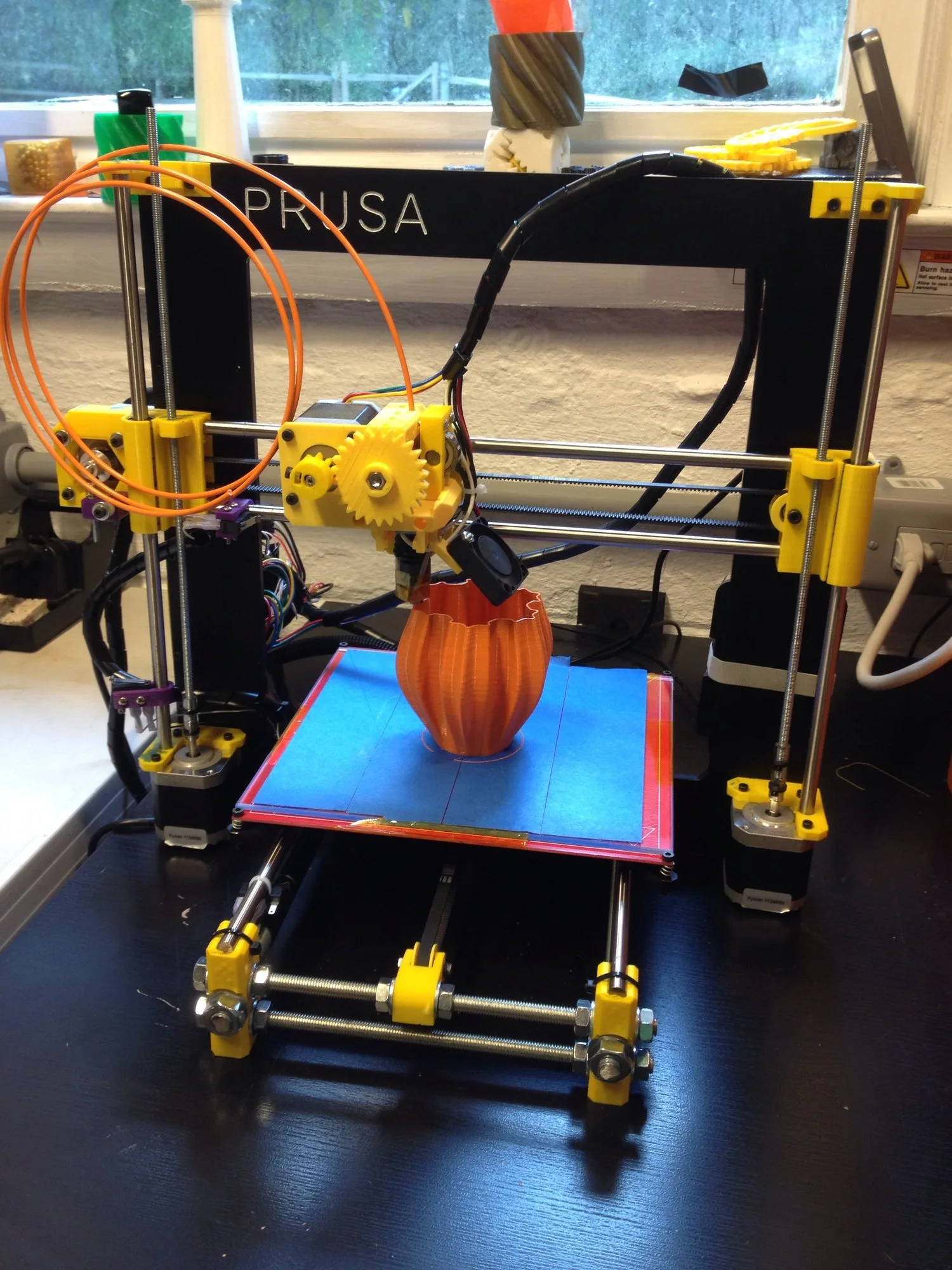 A Prusa i3 3D printer with yellow parts and orange plastic thread, printing an orange vase on a blue bed.